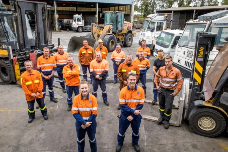 A team of Australian workers in hi-vis orange and navy workwear standing among forklifts, a front-end loader and trucks at an industrial worksite.