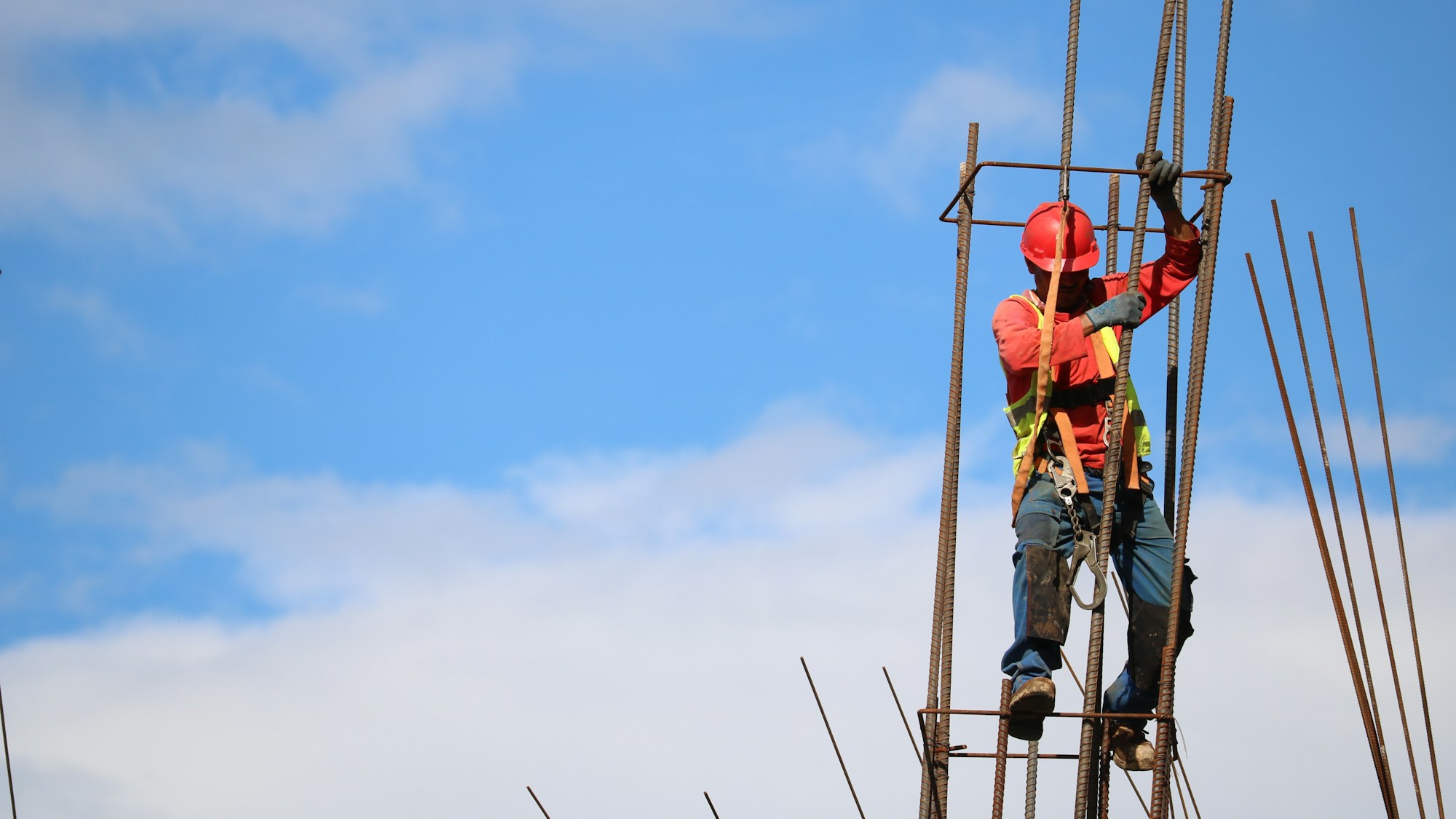 A construction worker in a red hard hat, hi-vis vest and safety harness climbing a steel reinforcing cage at height against a blue sky.